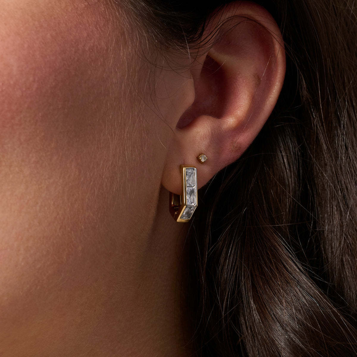 A close-up of a person's ear wearing a gold huggie hoop earring set with clear stones, alongside a small gold stud earring.
Gros plan sur l'oreille d'une personne portant une boucle d'oreille anneau dorée sertie de pierres transparentes, accompagnée d'une petite boucle d'oreille dorée.