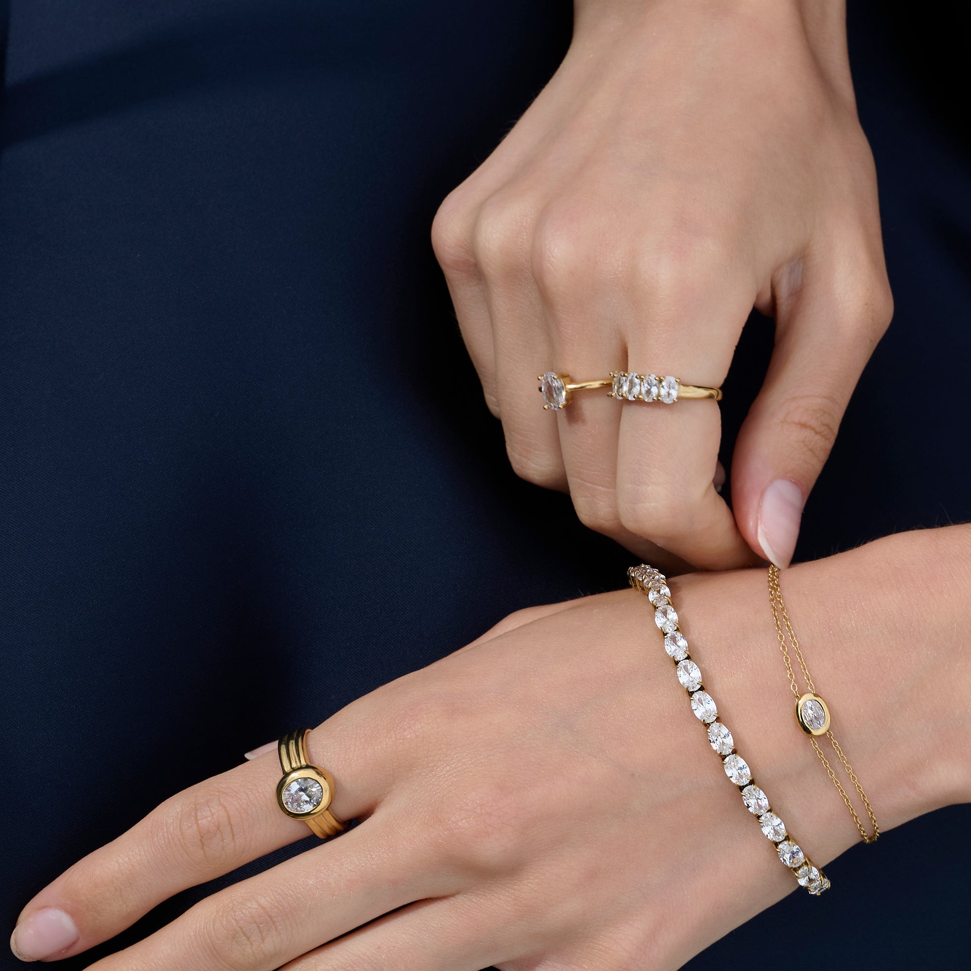 Close-up lifestyle shot of a woman's hands wearing a gold ring with a round clear stone and ridged band, paired with two stone-set gold rings and two gold bracelets against a navy background.
Photo lifestyle en gros plan des mains d'une femme portant une bague dorée sertie d'une pierre transparente ronde à anneau strié, agencée avec deux bagues dorées serties de pierres et deux bracelets dorés sur fond bleu marine.