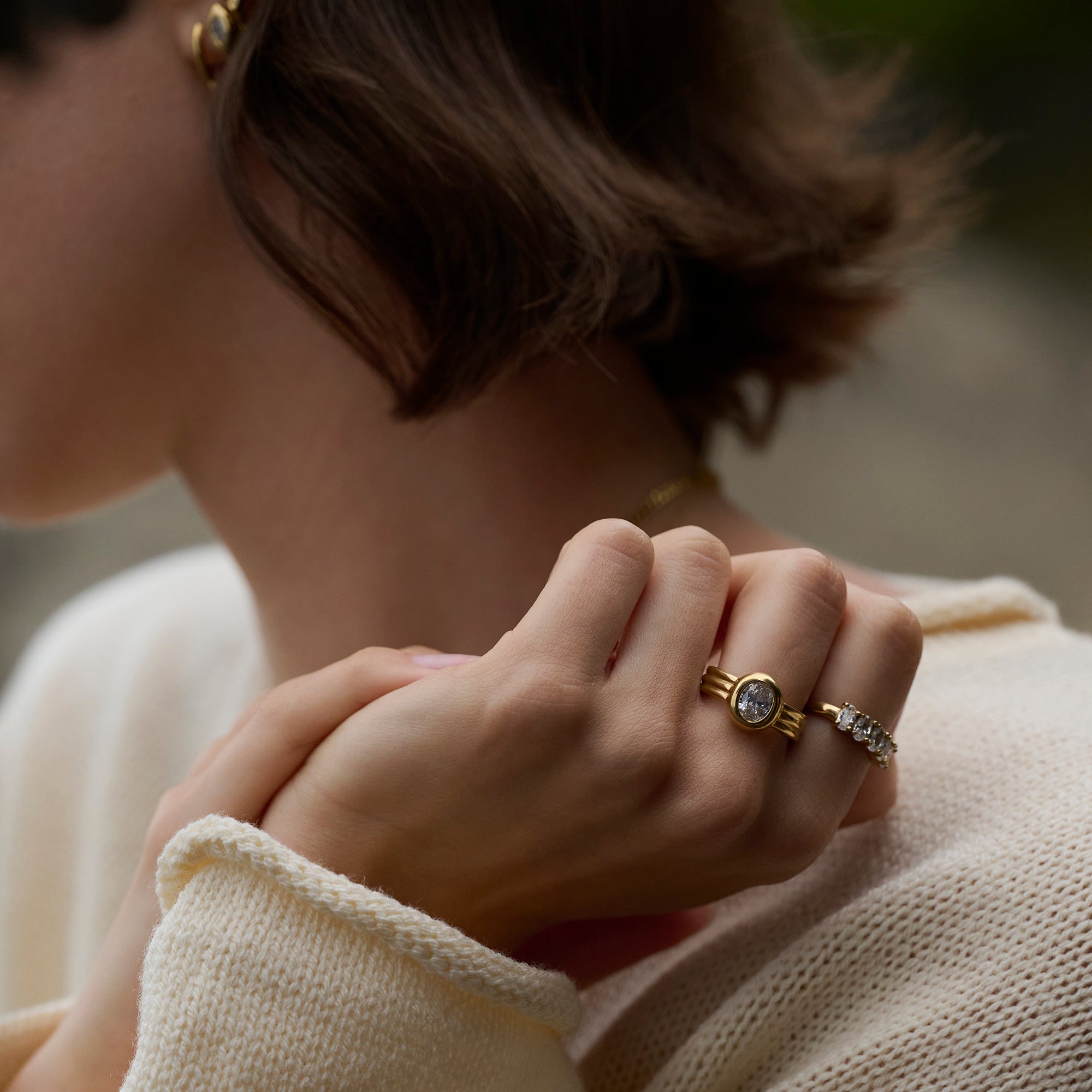 Close-up lifestyle shot of a woman's hands wearing a gold ring with a round clear stone and ridged band, paired with a stone-set gold ring and a cream knit sweater.
Photo lifestyle en gros plan des mains d'une femme portant une bague dorée sertie d'une pierre transparente ronde à anneau strié, agencée avec une bague dorée sertie de pierres et un tricot crème.