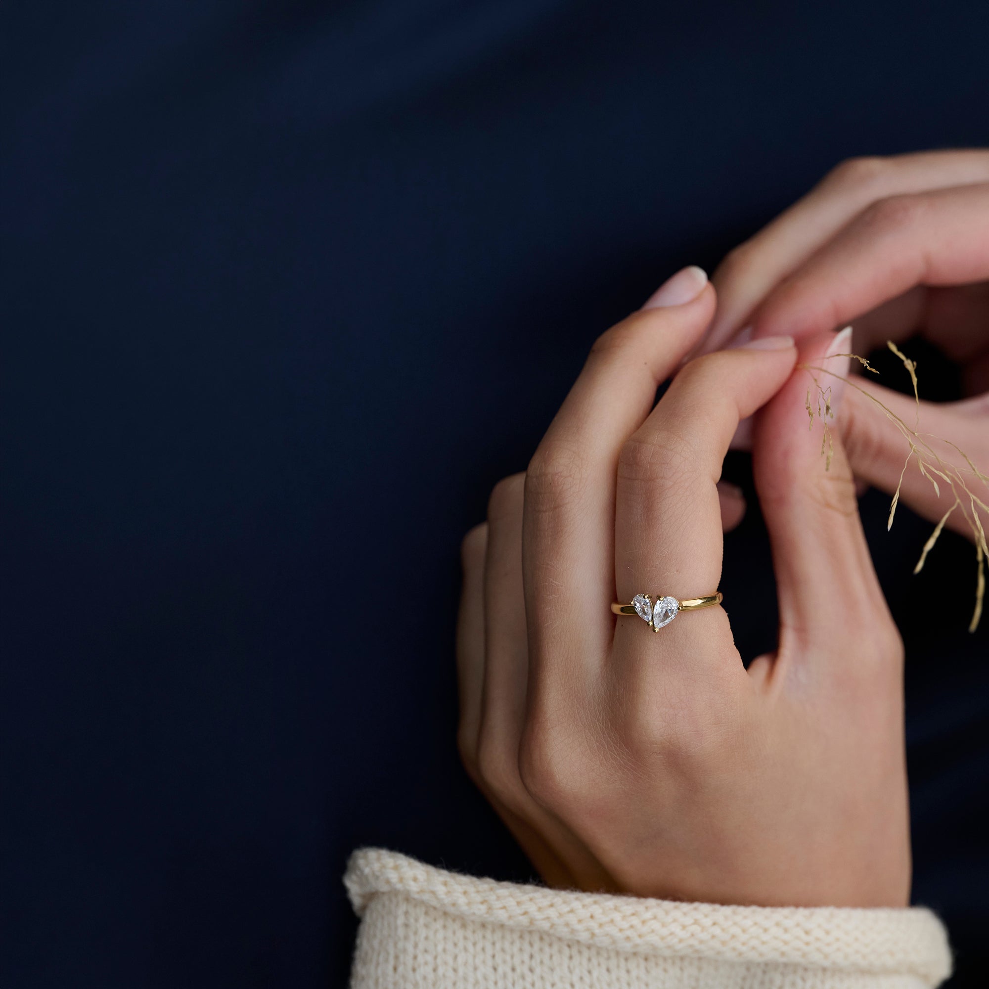 Close-up of a model's hands wearing a slim gold ring set with two pear-cut clear stones, styled with a cream knit sleeve against a dark navy background. Nickel-free, waterproof & tarnish-resistant.
Gros plan sur les mains d'un mannequin portant une bague fine dorée sertie de deux pierres transparentes taille poire, agencée avec une manche en tricot crème sur fond bleu marine foncé. Bague en acier inoxydable sans nickel, résistante à l'eau et anti-ternissement.