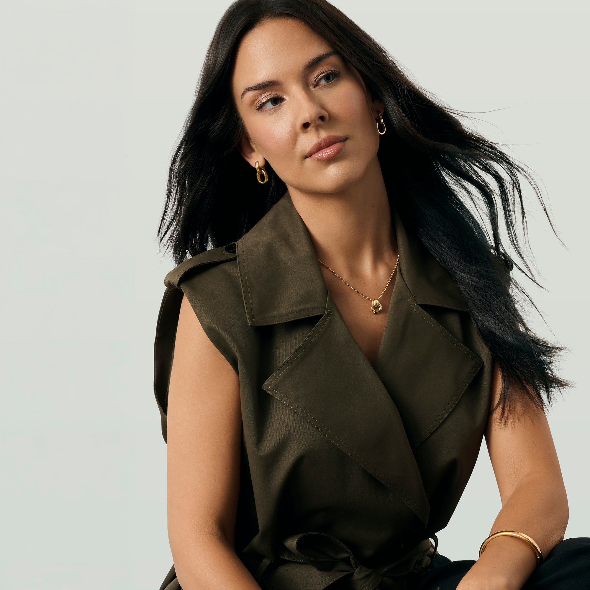 A woman wearing a sleeveless olive green top with a belted waist, styled with gold jewelry against a light gray studio background.
