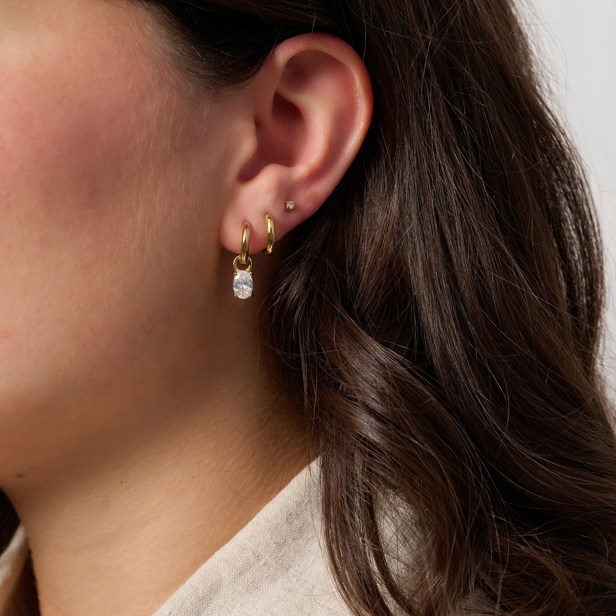 A close-up of a model's ear showcasing a gold huggie hoop earring with a dangling clear faceted gemstone, alongside other small earrings.