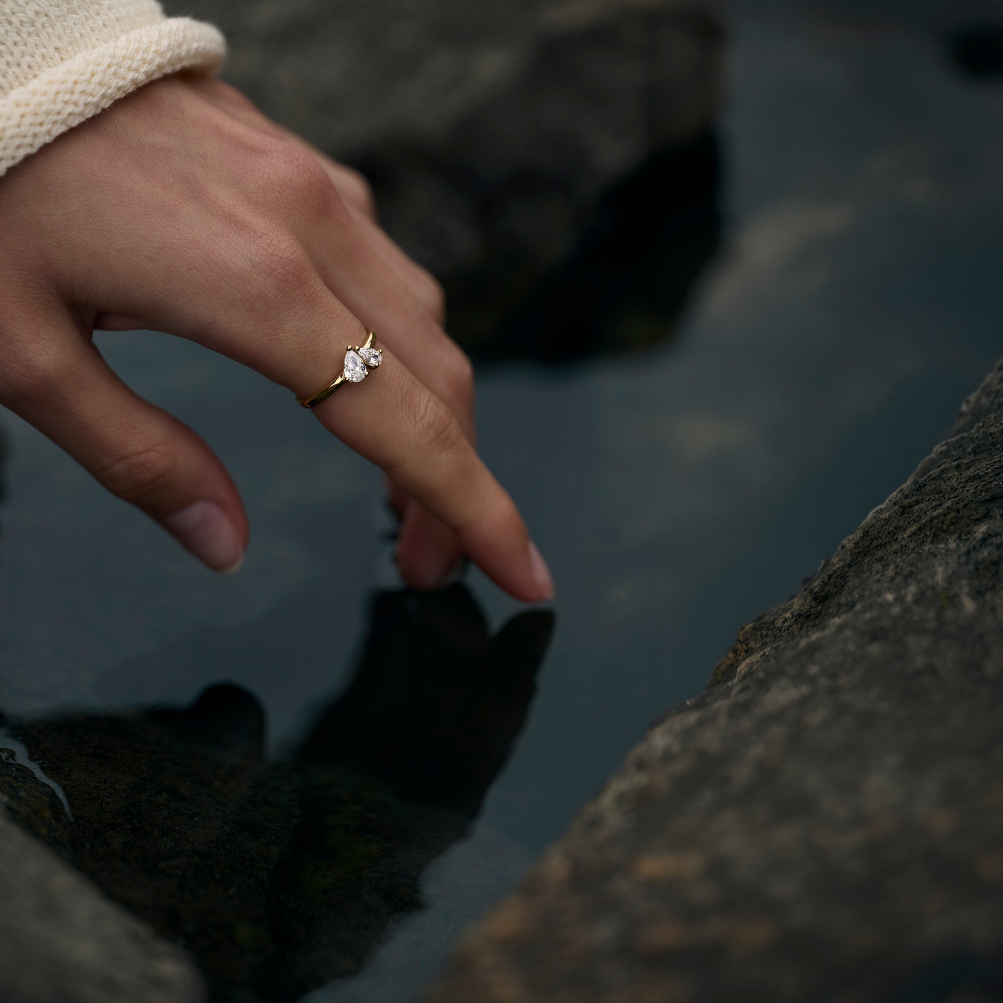 A close-up lifestyle shot of a hand wearing a delicate gold ring with a small clear gemstone, resting near dark water and rocks.
Photo lifestyle en gros plan d'une main portant une bague fine dorée sertie d'une petite pierre transparente, posée près d'une eau sombre et de rochers.