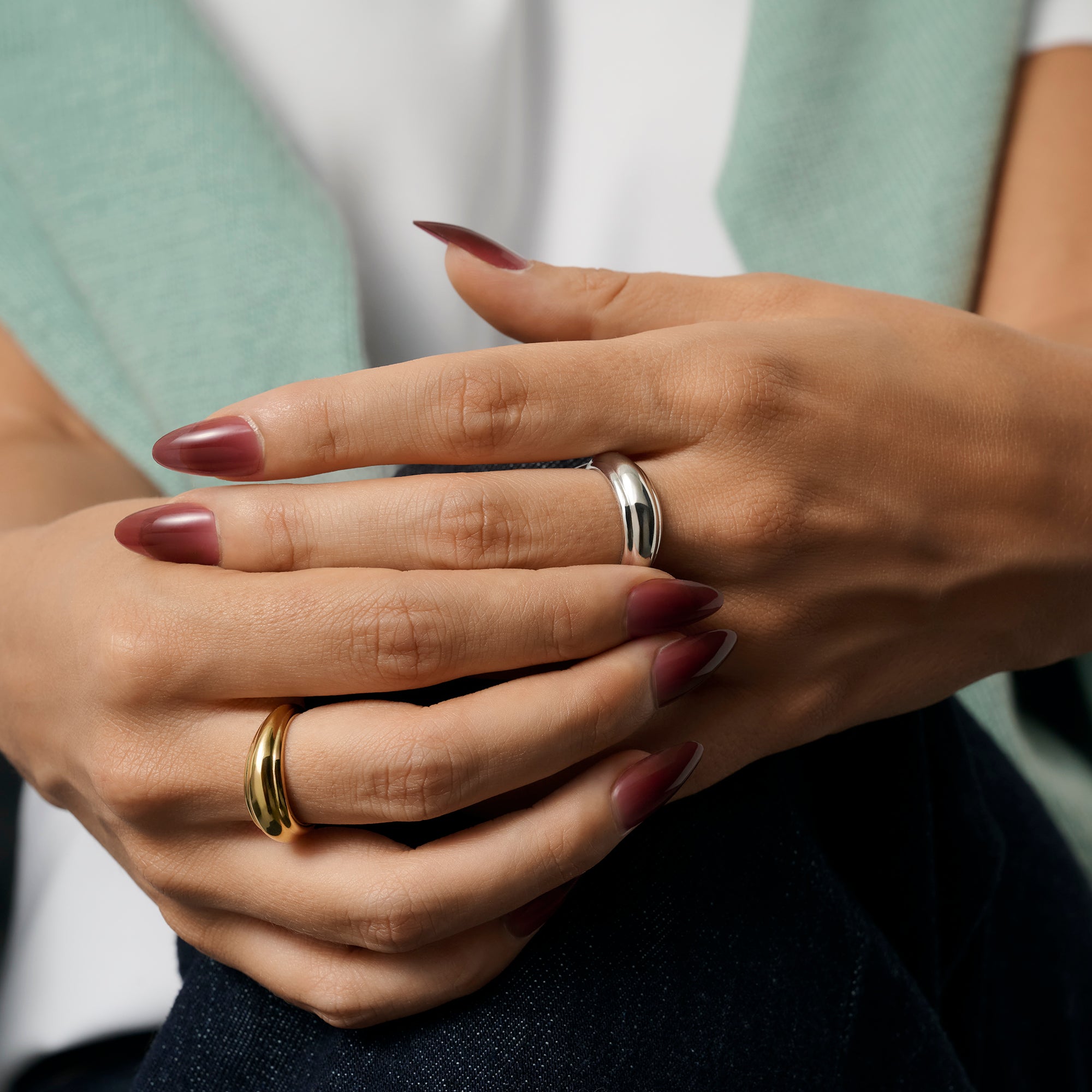 Close-up of a person's hands with dark burgundy manicured nails, wearing two curved dome rings in gold and silver, against a soft mint green and white top. Nickel-free, waterproof & tarnish-resistant stainless steel rings.
Gros plan sur les mains d'une personne aux ongles manucurés bourgogne foncé, portant deux bagues dôme galbées, l'une dorée et l'autre argentée, sur un haut vert menthe et blanc. Bagues en acier inoxydable sans nickel.