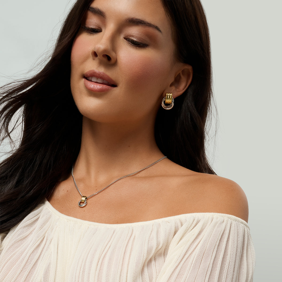 A close-up of a woman wearing a delicate two-tone necklace and matching earrings, paired with an off-the-shoulder cream top against a light gray background.
Gros plan d'une femme portant un collier délicat deux tons et des boucles d'oreilles assorties, agencés avec un haut crème à épaules dénudées sur fond gris pâle.