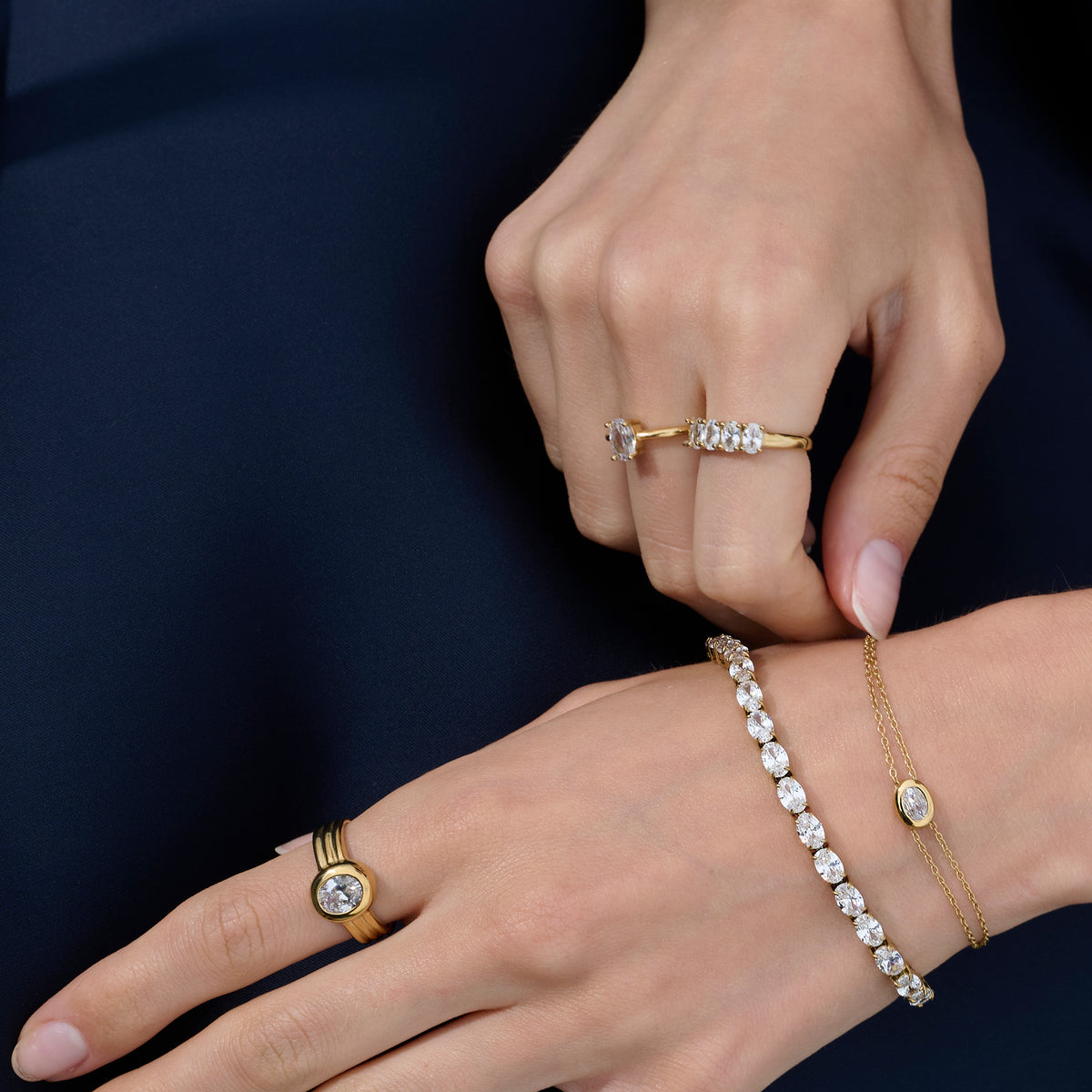 Close-up lifestyle shot of a woman's hands wearing a gold ring with a round clear stone and ridged band, paired with two stone-set gold rings and two gold bracelets against a navy background.

Photo lifestyle en gros plan des mains d'une femme portant une bague dorée sertie d'une pierre transparente ronde à anneau strié, agencée avec deux bagues dorées serties de pierres et deux bracelets dorés sur fond bleu marine.