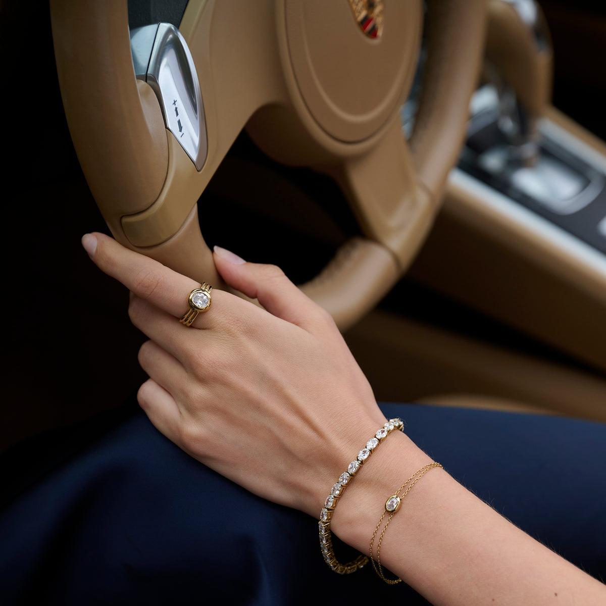 Close-up lifestyle shot of a woman's hand on a car steering wheel, wearing a gold ring with a round clear stone and ridged band, paired with a tennis bracelet and a fine chain bracelet. 

Photo lifestyle en gros plan de la main d'une femme sur un volant, portant une bague dorée sertie d'une pierre transparente ronde à anneau strié, agencée avec un bracelet tennis et un bracelet à chaîne fine.