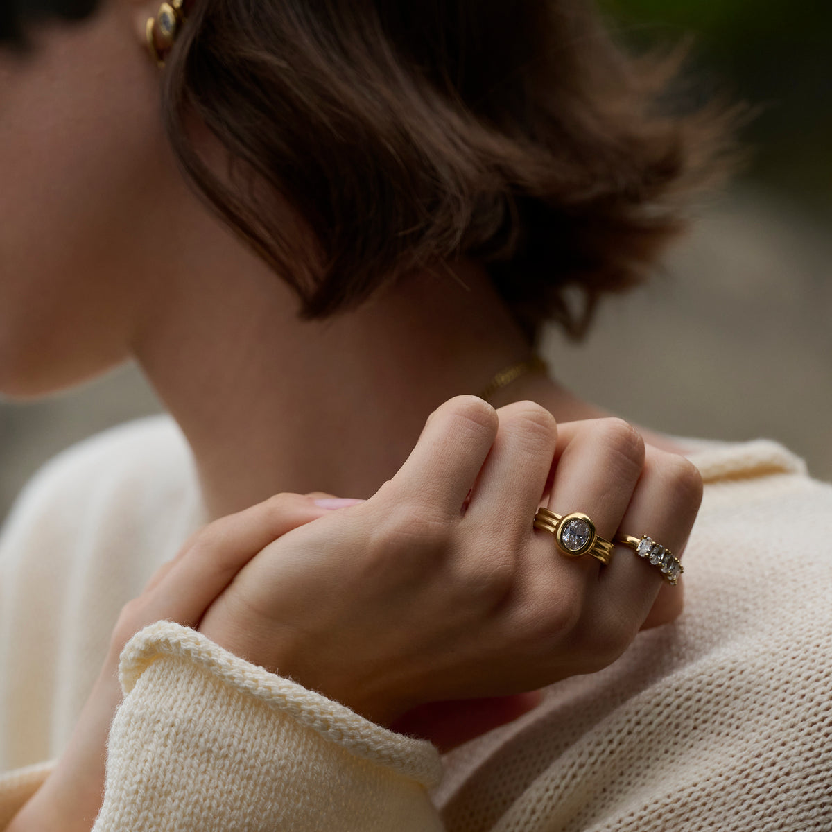 Close-up lifestyle shot of a woman's hands wearing a gold ring with a round clear stone and ridged band, paired with a stone-set gold ring and a cream knit sweater.

Photo lifestyle en gros plan des mains d'une femme portant une bague dorée sertie d'une pierre transparente ronde à anneau strié, agencée avec une bague dorée sertie de pierres et un tricot crème.