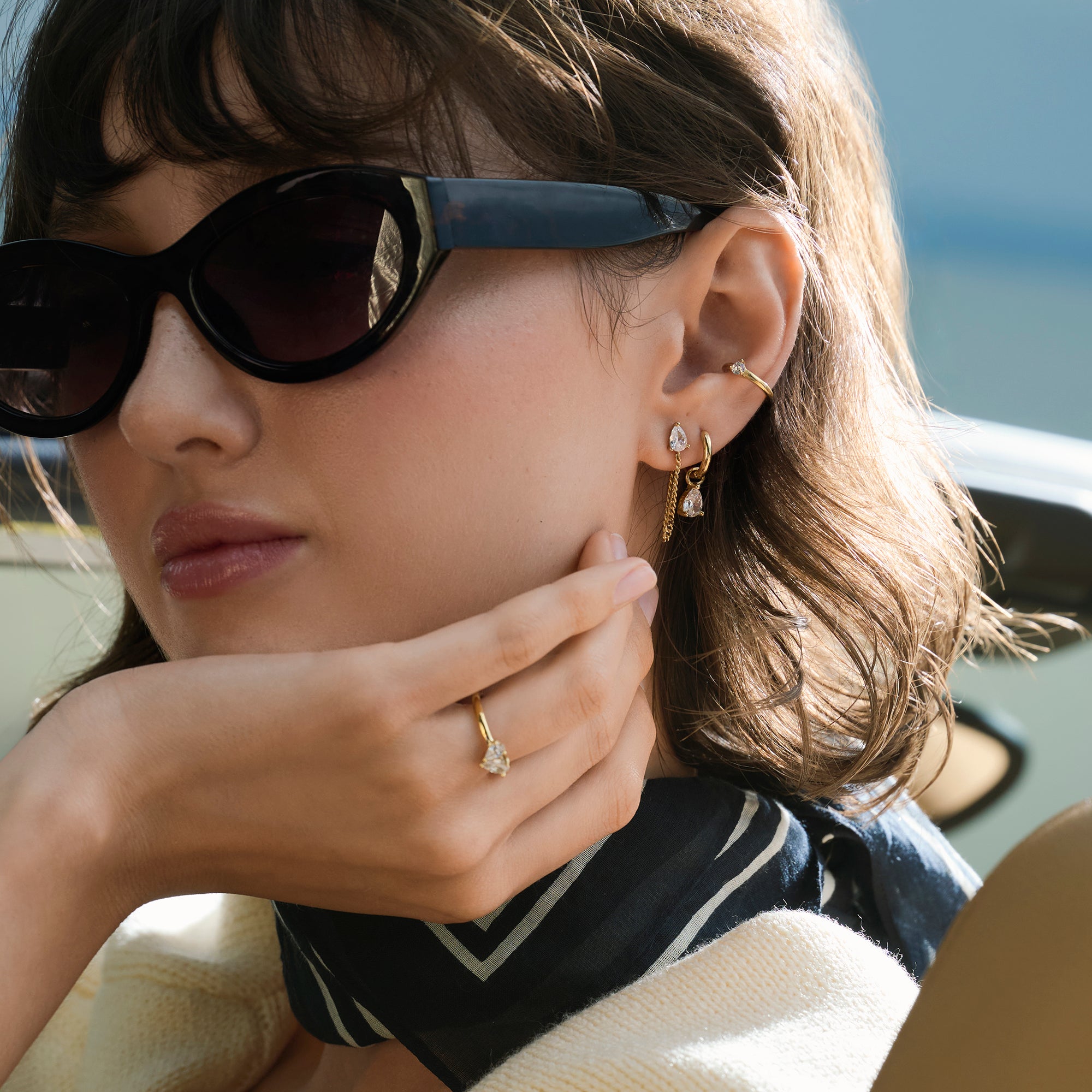 A close-up lifestyle shot of a woman wearing a gold pear-cut stone ring with matching gold earrings and ear cuff, styled with sunglasses, a navy scarf and a cream knit.

Photo lifestyle en gros plan d'une femme portant une bague dorée sertie d'une pierre taille poire, agencée avec des boucles d'oreilles dorées assorties et une manchette d'oreille, des lunettes de soleil, un foulard bleu marine et un tricot crème.