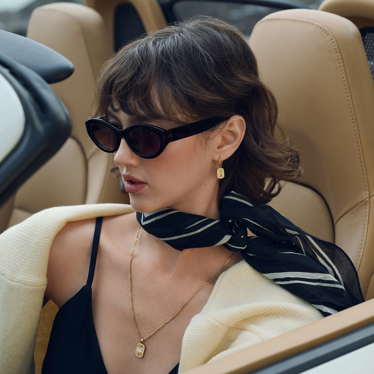 Close-up lifestyle shot of a woman seated in a car, wearing gold hoop earrings with a rectangular sunburst pendant and a matching gold chain necklace, styled with sunglasses, a striped scarf and a cream knit.

Photo lifestyle en gros plan d'une femme assise dans une voiture, portant des boucles d'oreilles anneaux dorées à pendentif rectangulaire rayonnant et un collier à chaîne dorée assorti, agencés avec des lunettes de soleil, un foulard rayé et un tricot crème.
