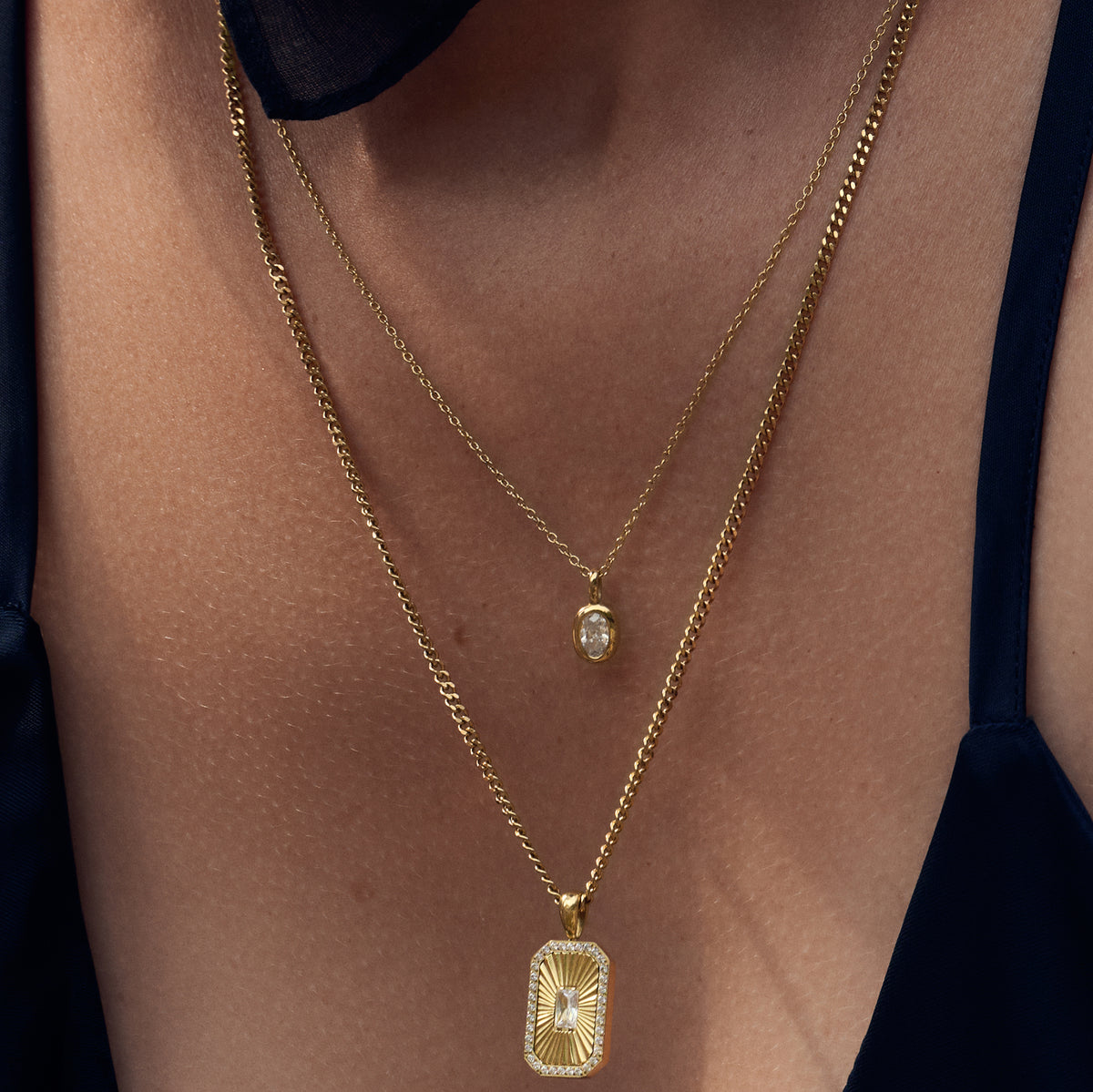 Close-up of a model wearing two layered gold necklaces, one with a small oval stone pendant and the other with a rectangular sunburst pendant. Waterproof, stainless steel and allergy free. By MIAJWL.

Gros plan d'un mannequin portant deux colliers dorés superposés, l'un à petit pendentif ovale serti et l'autre à pendentif rectangulaire rayonnant. Résistant à l'eau, en acier inoxydable et hypoallergénique. Par Mia Bijoux.